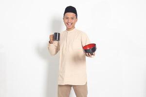 Portrait of excited Asian muslim man in koko shirt with skullcap holding a mug and empty bowl. Bowl template for food brand. Isolated image on white background photo