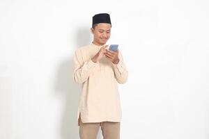 Portrait of young excited Asian muslim man in koko shirt with peci holding mobile phone with smiling expression on face. Social media and internet concept. Isolated image on white background photo