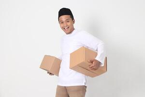 Portrait of attractive Asian muslim man in koko shirt with peci feeling grateful and peacful while carrying cardboard box. Going home for Eid Mubarak. Isolated image on white background photo