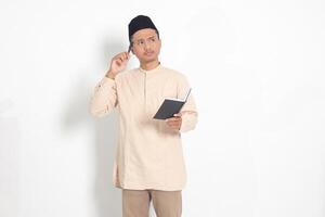 retrato de confuso asiático musulmán hombre en koko camisa con peci leyendo un libro, pensando acerca de un idea con mano participación bolígrafo en mentón, mirando lejos. aislado imagen en blanco antecedentes foto