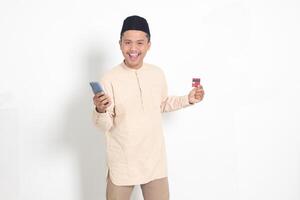 Portrait of attractive Asian muslim man in koko shirt with skullcap holding a mobile phone and presenting credit card. Isolated image on white background photo