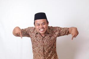 Portrait of excited Asian man wearing batik shirt and songkok smiling and looking at the camera pointing with two hands and fingers to the down side. Isolated image on gray background photo