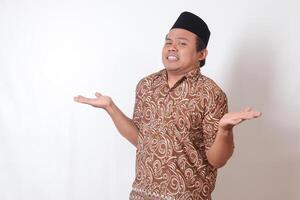 Portrait of confused Asian man wearing batik shirt and songkok with crossed hands, pointing sideways, making choice, choosing between two objects. Isolated image on gray background photo