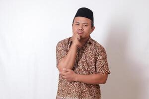 Portrait of confused Asian man wearing batik shirt and songkok standing against gray background, thinking about question with hand on chin photo