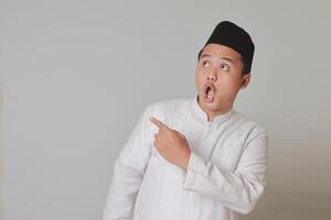 Portrait of Asian muslim man in white koko shirt poiting to the side and showing wow face expression. Isolated image on gray background photo