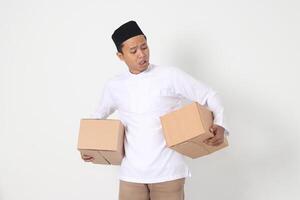 retrato de sorprendido asiático musulmán hombre en koko camisa con peci que lleva cartulina caja y haciendo conmocionado expresión. yendo hogar para eid mubarak. aislado imagen en blanco antecedentes foto