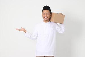 Portrait of attractive Asian muslim man in koko shirt with peci pointing to the side while carrying cardboard box. Going home for Eid Mubarak. Isolated image on white background photo