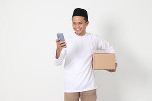 Portrait of excited Asian muslim man carrying cardboard box while holding mobile phone showing cheerful expression. Going home for Eid Mubarak. Isolated image on white background photo