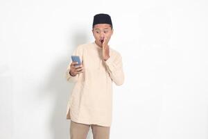 Portrait of surprised Asian muslim man in koko shirt with skullcap holding mobile phone, showing shocked face expression. Advertising and social media concept. Isolated image on white background photo