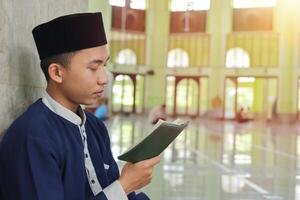 Religious Asian man in muslim shirt and black cap reading the holy book of Quran in the public mosque photo