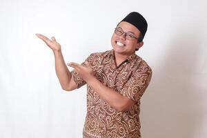 Portrait of excited Asian man wearing batik shirt and songkok smiling and looking at the camera pointing with two hands and fingers to the side. Isolated image on gray background photo