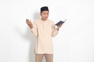 Portrait of confused Asian muslim man in koko shirt with peci difficulty understanding the contents of the book, reading a textbook. Isolated image on white background photo