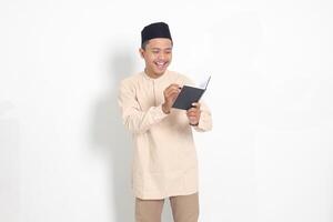 Portrait of excited muslim man in koko shirt with peci writing on his notebook. Happy Asian guy taking notes. Isolated image on white background photo