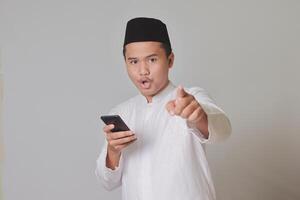 Portrait of Asian muslim man in white koko shirt with skullcap pointing forward to camera with his finger while using mobile phone. Isolated on gray background photo