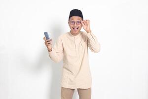 Portrait of surprised Asian muslim man in koko shirt with skullcap holding mobile phone, showing wow shocked face expression. Advertising and social media concept. Isolated image on white background photo