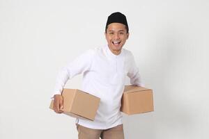 Portrait of happy Asian muslim man in koko shirt with peci looking confident while carrying cardboard box. Homecoming on Eid Mubarak concept. Isolated image on white background photo