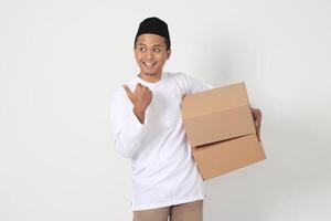 Portrait of excited Asian muslim man in koko shirt with peci carrying cardboard box while pointing and showing thumb up hand gesture. Going home for Eid Mubarak. Isolated image on white background photo