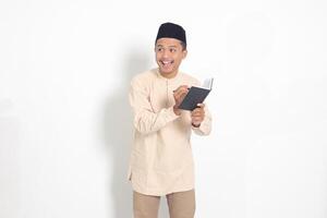 Portrait of excited muslim man in koko shirt with peci writing on his notebook. Happy Asian guy taking notes. Isolated image on white background photo