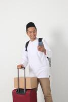 Portrait of happy Asian muslim man carrying cardboard box and suitcase while holding mobile phone. Going home for Eid Mubarak. Isolated image on white background photo