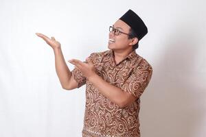 Portrait of excited Asian man wearing batik shirt and songkok smiling and looking at the camera pointing with two hands and fingers to the side. Isolated image on gray background photo