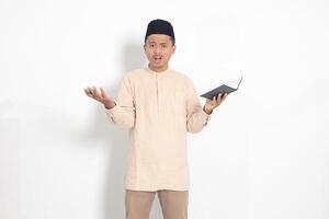 Portrait of confused Asian muslim man in koko shirt with peci difficulty understanding the contents of the book, reading a textbook. Isolated image on white background photo