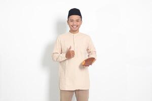 Portrait of excited Asian muslim man in koko shirt with skullcap showing and holding an empty plate. Bowl template for food brand. Isolated on white background photo