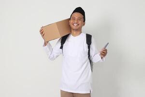 Portrait of happy Asian muslim man holding mobile phone while carrying cardboard box with excited expression. Going home for Eid Mubarak. Isolated image on white background photo