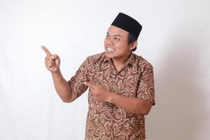 Portrait of excited Asian man wearing batik shirt and songkok smiling and looking at the camera pointing with two hands and fingers to the side. Isolated image on gray background photo