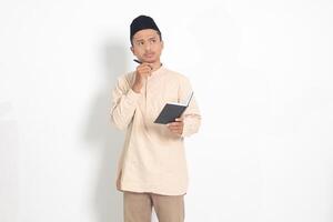 Portrait of confused Asian muslim man in koko shirt with peci reading a book, thinking about an idea with hand holding pen on chin, looking away. Isolated image on white background photo