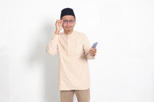 Portrait of surprised Asian muslim man in koko shirt with skullcap holding mobile phone, showing wow shocked face expression. Advertising and social media concept. Isolated image on white background photo