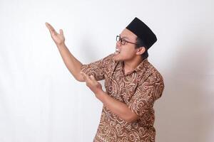 Portrait of excited Asian man wearing batik shirt and songkok smiling and looking at the camera pointing with two hands and fingers to the side. Isolated image on gray background photo