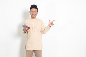 Portrait of attractive Asian muslim man in koko shirt with skullcap holding a mobile phone and credit card while pointing with finger to the side. Isolated image on white background photo