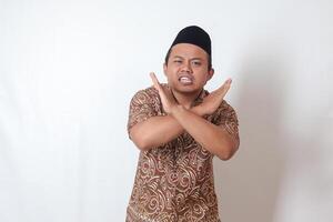 Portrait of excited Asian man wearing batik shirt and songkok  showing X sign of hands, refusing an invitation from someone, making prohibitive gesture. Isolated image on gray background photo
