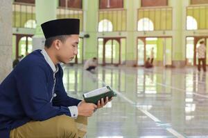 Religious Asian man in muslim shirt and black cap reading the holy book of Quran in the public mosque photo