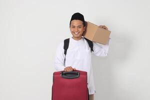 Portrait of excited Asian muslim man in koko shirt with peci carrying cardboard box and holding suitcase. Going home for Eid Mubarak. Isolated image on white background photo