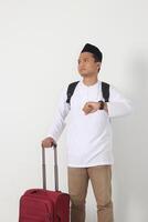 Portrait of displeased Asian muslim man standing over white background, holding the suitcase handle, paying attention to the time on watch preparing for the trip. Going home for Eid Mubarak. photo