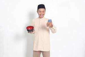 retrato de emocionado asiático musulmán hombre en koko camisa con casquete participación un móvil teléfono y vacío bol. cuenco modelo para comida marca. social medios de comunicación publicidad concepto. aislado en blanco antecedentes foto