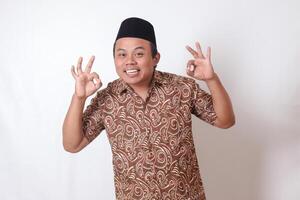 Portrait of excited Asian man wearing batik shirt and songkok showing ok hand gesture and smiling looking at camera. Isolated image on gray background photo