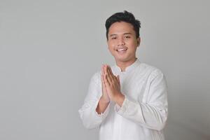 Portrait of Asian muslim man in white koko shirt showing apologize and welcome hand gesture. Isolated image on gray background photo
