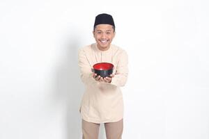 Portrait of excited Asian muslim man in koko shirt with skullcap showing and holding an empty plate. Bowl template for food brand. Isolated on white background photo