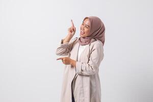 Portrait of excited Asian hijab woman in casual suit pointing and showing product in her side with finger. Businesswoman concept. Isolated image on white background photo