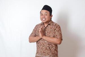 Portrait of smiling Asian man wearing batik shirt and songkok showing apologize and welcome hand gesture. Ramadan advertising concept. Isolated image on gray background photo