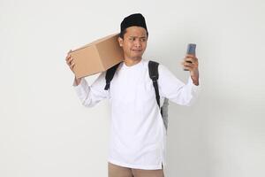 Portrait of worried Asian muslim carrying cardboard box while having having call on mobile phone, showing sad expression. Going home for Eid Mubarak. Isolated image on white background photo