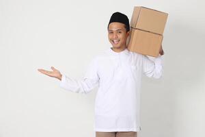 Portrait of excited Asian muslim man in koko shirt with peci carrying cardboard box while pointing to the side. Going home for Eid Mubarak. Isolated image on white background photo