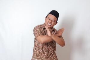 retrato de emocionado asiático hombre vistiendo batik camisa y Songkok demostración X firmar de manos, negarse un invitación desde alguien, haciendo prohibitivo gesto. aislado imagen en gris antecedentes foto