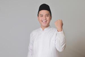 retrato de asiático musulmán hombre en blanco koko camisa con casquete levantamiento su puño, celebrando éxito. aislado imagen en gris antecedentes foto