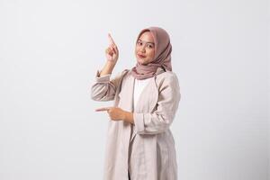 Portrait of excited Asian hijab woman in casual suit pointing and showing product in her side with finger. Businesswoman concept. Isolated image on white background photo