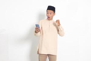 Portrait of excited Asian muslim man in koko shirt with skullcap raising his fist, celebrating winning game on mobile phone. Advertising and social media concept. Isolated image on white background photo