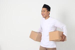 Portrait of happy Asian muslim man in koko shirt with peci looking confident while carrying cardboard box. Homecoming on Eid Mubarak concept. Isolated image on white background photo