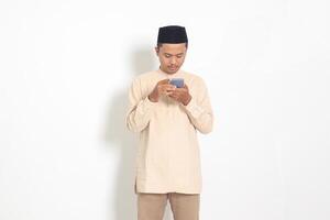 Portrait of young excited Asian muslim man in koko shirt with peci holding mobile phone with smiling expression on face. Social media and internet concept. Isolated image on white background photo